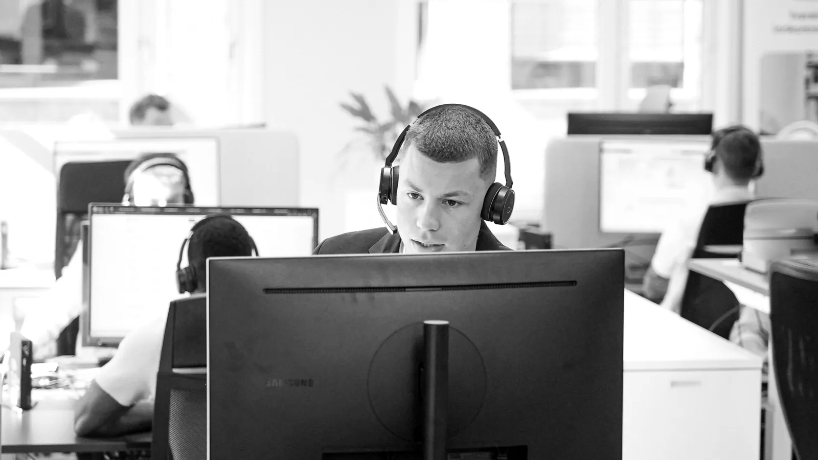 Selger med headset ved skjermen, med flere kolleger i arbeid på salgsgulvet bak